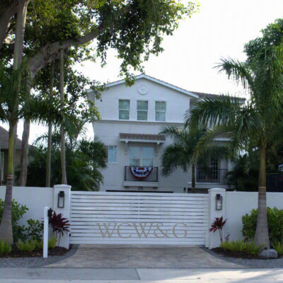 White two-story house with a gated entrance, palm trees, and a patriotic banner hanging on the balcony railing.