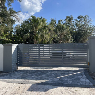Gray metal sliding gate between two pillars, with trees and a blue sky in the background.