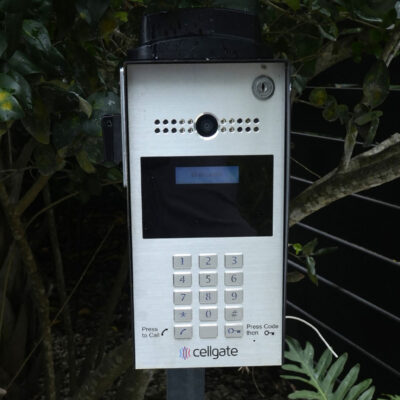 A Cellgate intercom system with keypad and camera is installed outdoors near plants.