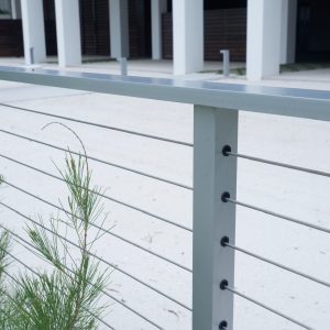 Metal railing with horizontal cables and nearby green plant, with a white building and paved area in the background.
