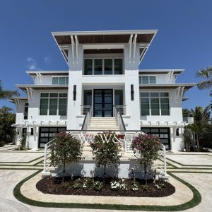 Modern three-story white house with large windows, front steps, and landscaped garden, photographed on a sunny day.