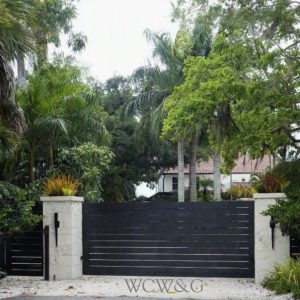 Black horizontal gate with stone pillars and dense tropical greenery, partially obscuring a house in the background.