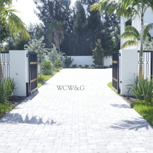 Paved driveway lined with plants and palm trees, leading to a modern house gate with open black iron gates.