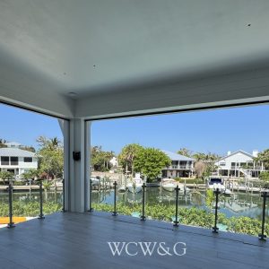 Spacious room with open glass walls overlooks a canal lined with houses, boats, trees, and blue sky.