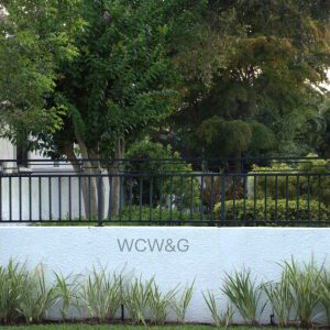 A white wall with “WCW&G” text, black metal fence, trees, and greenery behind; shrubs planted in front.