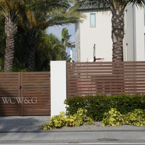 Modern wooden gate with “WCW&G” lettering, flanked by white walls, palm trees, and shrubs in front of a contemporary house.