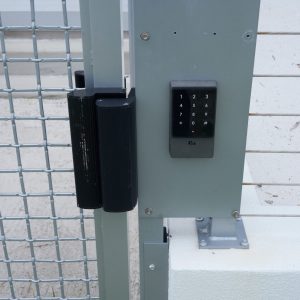 Close-up of a metal gate with an electronic keypad lock and wire mesh panel beside a white concrete wall.