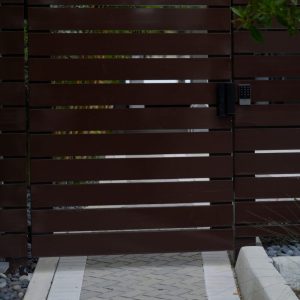 Brown horizontal wooden gate with a keypad lock, positioned at the end of a paved walkway bordered by rocks.