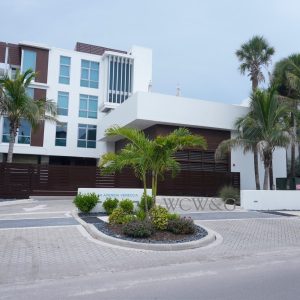 Modern white apartment building with balconies, surrounded by palm trees and landscaped driveway, under a cloudy sky.
