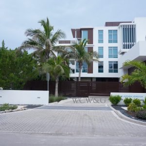 Modern three-story building with palm trees, white walls, brown accents, and landscaped driveway under a cloudy sky.