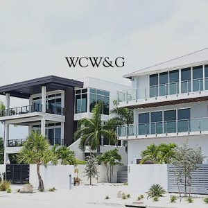 Modern multi-story beach houses with large windows and balconies, surrounded by palm trees under a cloudy sky.