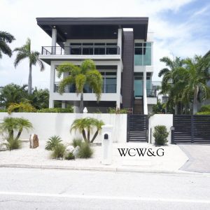 Modern three-story house with large windows, palm trees, white exterior, and black gate on a sunny day.