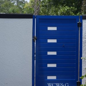 A blue gate with rectangular cutouts and the letters “WCW&G” at the bottom, set in a white wall.