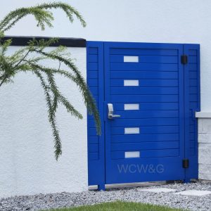A modern blue metal gate with rectangular glass panels stands next to a white wall and a small tree.