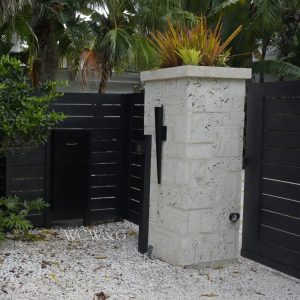 Modern black gate and mailbox next to a stone pillar with a plant on top, surrounded by tropical greenery.