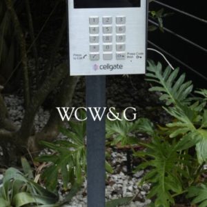 A silver cellgate intercom keypad mounted on a post, surrounded by plants and white gravel.
