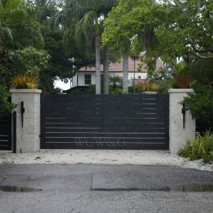 Large modern black gate with horizontal slats blocks the driveway entrance to a house surrounded by lush greenery.