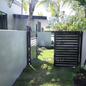 A modern black metal gate is open between white walls, leading to a grassy yard with palm trees.