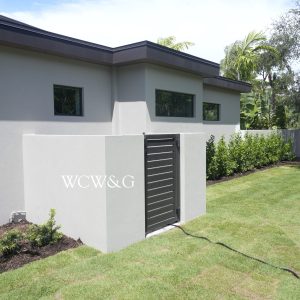 Modern house exterior with gray walls, black metal gate, manicured lawn, and surrounding greenery on a sunny day.