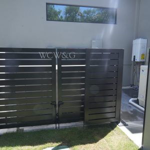 Black metal double gate partially open, revealing pool equipment and water heater beside a light gray building wall.