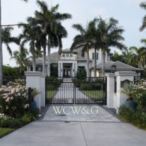 Gated entrance to a large, modern mansion with palm trees, manicured lawns, and flower beds under a cloudy sky.