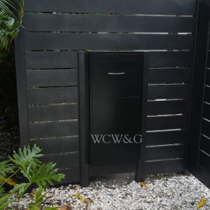 Black mailbox set into a slatted black wooden fence, surrounded by tropical plants and white gravel ground.