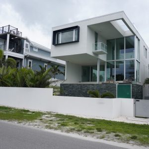 Modern white house with large glass windows next to a gray house, both surrounded by palm trees and white fencing.