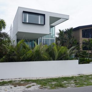Modern two-story white house with large windows, surrounded by palm trees and a white wall along the street.