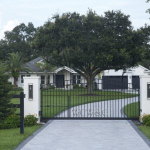 Black metal gate with stone pillars and a curved driveway leading to a white house with a large tree in front.