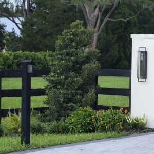 Black metal gate and fence beside landscaped greenery, flowers, and a white pillar with an outdoor light fixture.