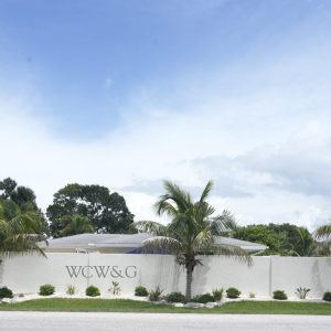 White wall with “WCW&G” text, three palm trees, and small shrubs in front; houses and trees visible behind the wall.