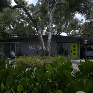 Single-story house with gray walls, neon green doors, a carport, and large trees in front, seen behind green shrubs.