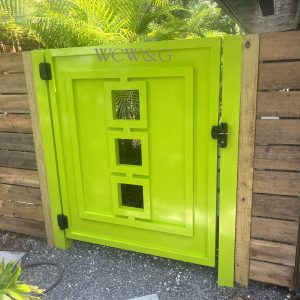 Bright green wooden gate with square cutout windows, attached to a wooden fence, surrounded by plants.