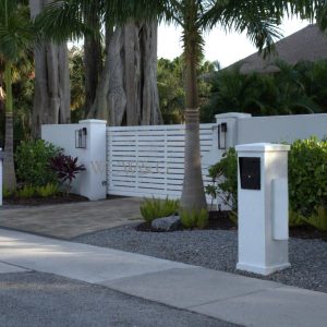 White gated entrance to a residential property, surrounded by palm trees and green landscaping beside a quiet street.