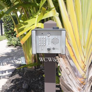 A metal intercom keypad stands by a sidewalk next to a large plant and a street.