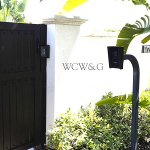 Black gate with keypad and speaker post next to a white wall displaying the letters “WCW&G” and greenery below.