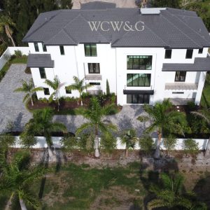 Aerial view of a large white modern house with a fenced yard, palm trees, and a driveway.