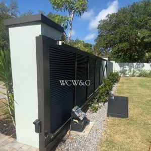 Black metal gate with slats, attached to a pale wall, next to a control box and a neatly maintained lawn.