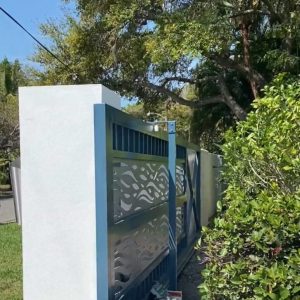 Blue metal gate with decorative cutouts, attached to a white wall, partially open near greenery and a street.