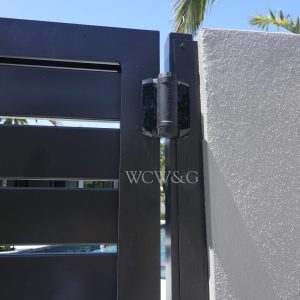 Close-up of a black metal gate latch attached to a white concrete wall, with palm trees in the background.