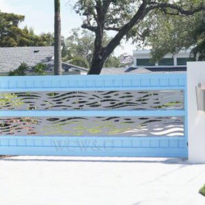 Light blue modern driveway gate with horizontal wave cutouts, set between white walls and surrounded by greenery.