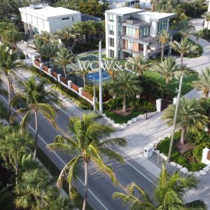A modern, multi-story house with large windows is surrounded by palm trees and a gated driveway in a tropical setting.