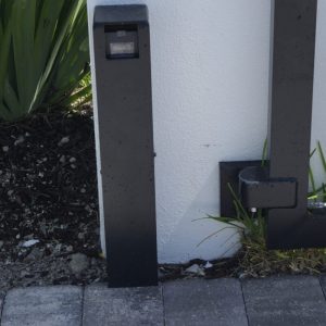 Black outdoor light post next to a white wall with “WCW&G” text, plants, and pavement in view.