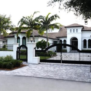 Large white mansion with palm trees, arched windows, and a black gated entrance on a paved driveway.