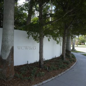 White wall with the letters “WCW&G” next to a sidewalk, shaded by trees and surrounded by landscaping.