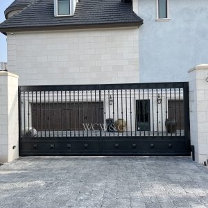 Black metal driveway gate with decorative details in front of a modern, light-colored house with a stone facade.