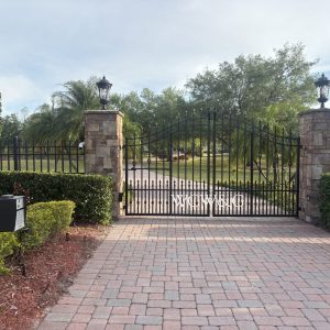 Black iron gate with stone pillars blocks entrance to a brick driveway lined with bushes and trees.