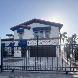 Two-story white house with blue awnings and a red tile roof, behind a decorative black metal gate and driveway.