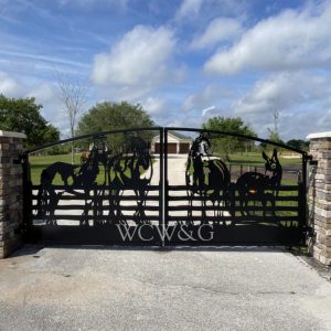 Metal gate with cutout silhouettes of horses, cowboys, and WCW&G initials, set between stone pillars on a ranch driveway.