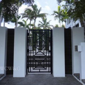 Black metal gate set in a white wall with palm trees and a modern house visible in the background under a partly cloudy sky.